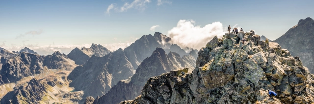 Felsiger Gipfel mit Menschen vor einer felsigen Bergkulisse unter trübem blauen Himmel mit Wolken