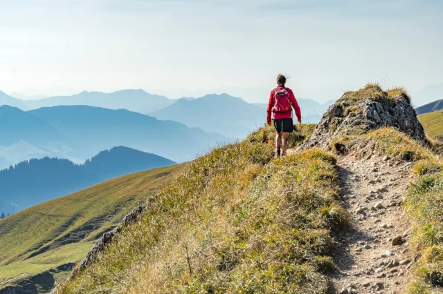 Frau mit Rucksack läuft auf einem leichten Grat mit Wiesen mit Bergen im Hintergrund