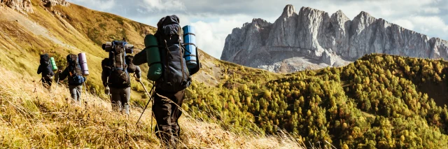 Eine Wandergruppe mit schwer bepackten Rucksäcken geht durch eine hohe Wiese mit Bergpanorama im Hintergrund. 