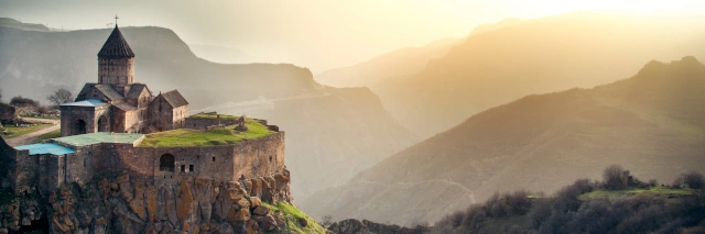 Blick auf das Tatev Kloster das direkt auf einen hohen Felsen gebaut ist und vor einer Berglandschaft liegt. Im Hintergrund geht die Sonne unter.