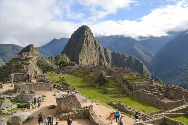 Blick auf die Ruinen von Maccu Picchu und den einzeln stehenden Felsen. Über der Hügelkette im Hintergrund hängen weiße Wolken. 