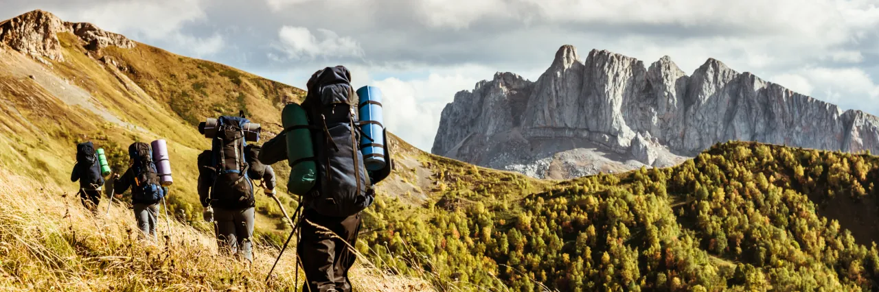 Eine Wandergruppe mit schwer bepackten Rucksäcken geht durch eine hohe Wiese mit Bergpanorama im Hintergrund. 