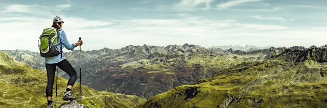 Eine Frau mit Rucksack und Wanderstöcken schaut auf eine Gebirgslandschaft hinunter. 