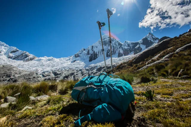 Ein blauer Wanderrucksack liegt neben zwei in den Boden gesteckten Wanderstöcken auf einer Bergwiese. Im Hintergrund ist eine Gebirgskette zu sehen, die mit Schnee bedeckt ist. 