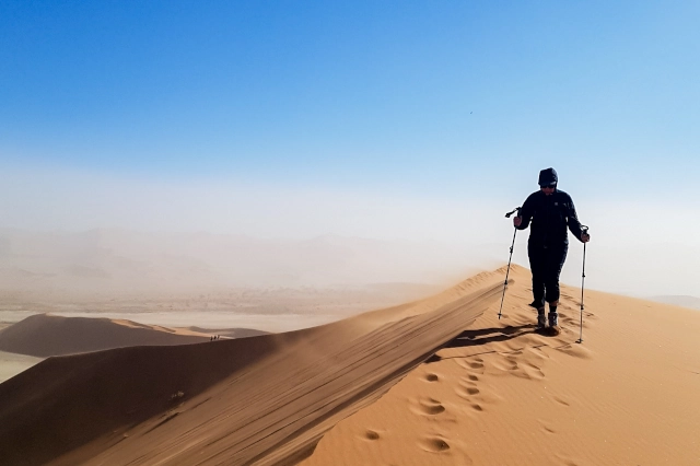 Eine Frau mit Trekkingstöcken folgt bereits vorhandenen Fußspuren auf dem Kamm einer einzelnen hohen Düne mit braunem Sand. Der Himmel ist strahlend blau. 
