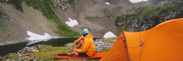 Ein Mann sitzt auf einer Isomatte vor seinem Zelt und schaut auf eine felsige Landschaft. 