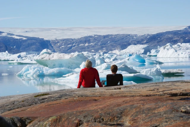 Ein Mann und eine Frau sitzen mit dem Rücken zur Kamera auf dem Boden. Sie blicken auf eine Wasseroberfläche mit Eisschollen. Im Hintergrund ist eine hügelige Landschaft zu sehen. 
