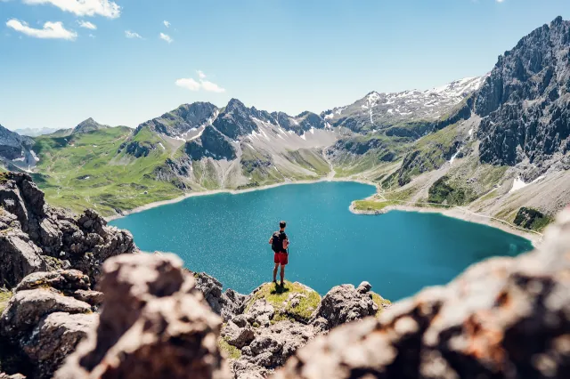 Mann mit Rucksack blickt über einen blauen Bergsee, der umrundet ist von kleineren Bergen