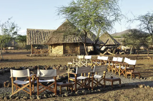 Um eine Feuerstelle stehen Stühle. Im Hintergrund sind mehrere traditionelle Gebäude zu sehen, die in savannenmäßiger Landschaft stehen. 