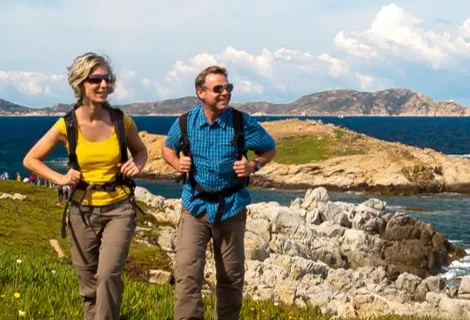 An einer Küste stehen eine Wanderin und ein Wanderer auf einer Wiese mit Löwenzahn.