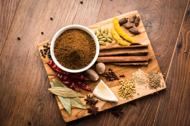 Auf einem Holzbrett liegen eine Vielzahl an verschiedenen Gewürzen, wie getrocknete Chilischoten, Zimtstangen, Sternanis und Nelken. Außerdem steht eine weiße Schüssel mit einem braunen Gewürzpulver. 