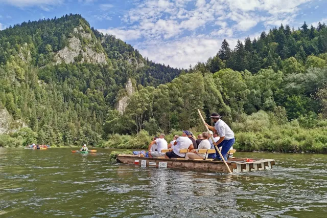 Eine kleine Gruppe sitzt auf einem hölzernen, stabilen Floß. Hinten steht ein Flößer mit einem Stock. Das Floß treibt auf einem grünblauen Fluss. Im Hintergrund ist Nadelwald zu sehen. 