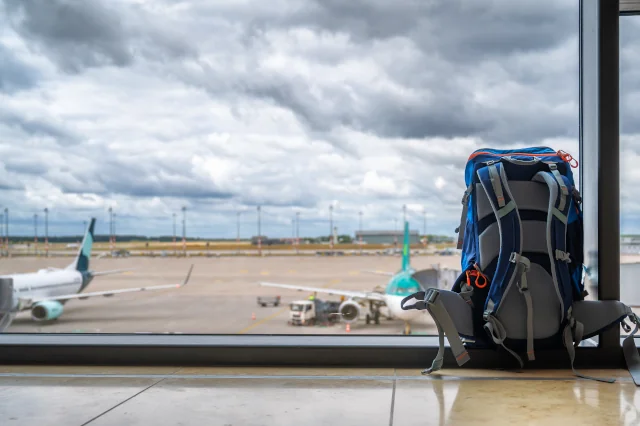 Ein Rucksack steht vor einem großen Fenster mit Blick auf das Außengelände des Flughafens München. 