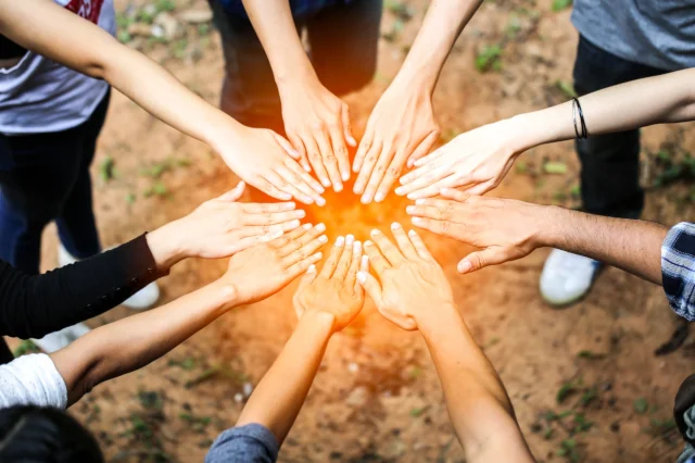 Eine Gruppe Menschen steht in einem Kreis und jeder streckt eine Hand in die Mitte. Die Hände ergeben einen Kreis, der leicht orange erleuchtet ist. 