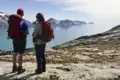 Zwei Wanderer mit Wanderstöcken und Rucksäcken stehen auf felsigem Gelände und blicken auf eine Bucht mit Bergen und Eisbergen.
