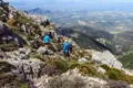 Zwei Wanderer auf einem Bergpfad, umgeben von Felsen und Vegetation, mit Blick auf ein Tal im Hintergrund.