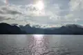 Sonnenlicht glitzert auf einem See, im Hintergrund sind bewaldete Berge und Wolkenformationen zu sehen.