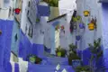 Eine Treppe führt eine enge Gasse in Chefchaouen hinauf, deren Wände in verschiedenen Blautönen gestrichen sind. Bunte Blumentöpfe hängen an den Wänden und stehen auf den Stufen.