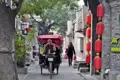 Rikscha-Fahrer transportiert Fahrgäste durch eine enge Gasse, vorbei an traditionellen Gebäuden und roten Laternen.