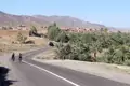Radfahrer auf einer Straße in der Nähe einer Oase und eines Dorfes in einer bergigen Wüstenlandschaft.