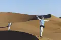 Zwei Reisende in einer sandigen Wüstenlandschaft. Eine Person steht mit ausgebreiteten Armen auf einer Düne.