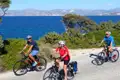 Drei Personen fahren mit Fahrrädern auf einer Küstenstraße. Im Hintergrund sind das Meer und Berge zu sehen.