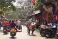 Straßenszene in Hanoi mit Fußgängern, Fahrradfahrern, Motorradfahrern und Händlern, die die geschäftige Atmosphäre der Stadt zeigen.