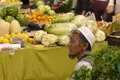 Mann an einem Marktstand mit Chinakohl, Blumenkohl, Zitronen, Melonen und weiterem Obst und Gemüse.