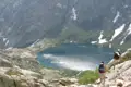 Zwei Wanderer auf einem Bergpfad. Ein Bergsee ist im Hintergrund zu sehen, umgeben von felsigen Hängen und Resten von Schneefeldern.