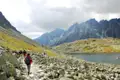 Wanderer auf einem steinigen Weg in den Bergen, mit einem Bergsee und Gipfeln im Hintergrund.