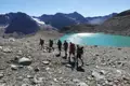 Wandergruppe auf einem Bergpfad neben einem türkisfarbenen See mit Bergen und Gletscher im Hintergrund.