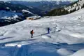 Zwei Schneeschuhwanderer auf einem verschneiten Bergpfad. Eine Hütte ist in der Ferne sichtbar, ebenso wie die umliegenden Berge.