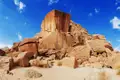 Eine große, rotbraune Felsformation ragt in den blauen Himmel. Am Fuß der Formation liegen weitere Felsen und Sand.