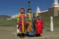 Drei Kinder in traditioneller mongolischer Kleidung vor einer Reihe von weißen Stupas in der Mongolei.