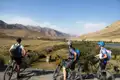 Drei Mountainbiker mit Rucksäcken und Helmen halten an einem Aussichtspunkt an und betrachten die Landschaft mit Fluss und Bergen.