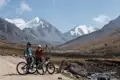 Zwei Personen auf Mountainbikes pausieren auf einem Bergpfad mit schneebedeckten Bergen im Hintergrund.