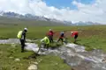 Mountainbiker schieben ihre Fahrräder durch einen Fluss in einer Gebirgslandschaft.