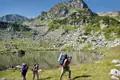 Drei Wanderer mit schweren Rucksäcken beim Trekking in den Bergen. Ein Bergsee und felsige Landschaft im Hintergrund.