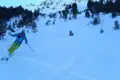 Skifahrer im Tiefschnee zwischen Bäumen mit schneebedeckten Bergen im Hintergrund.