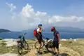 Zwei Radfahrer mit Helmen und Rucksäcken machen eine Pause an der Küste und trinken Wasser. Im Hintergrund ist das Meer und eine bergige Landschaft zu sehen.
