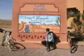 Radreise in Marokko: Pause in Zagora vor einem Schild mit der Aufschrift 'Timbuktu 52 Tage'.