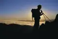 Silhouette eines Wanderers mit Rucksack und Wanderstöcken, der auf einem Berggipfel steht und den Sonnenaufgang über einem Wolkenmeer beobachtet.