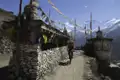 Radfahrer auf einem Bergpfad im Himalaya, vorbei an traditionellen Steinbauten mit Gebetsfahnen und schneebedeckten Bergen im Hintergrund.