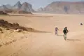 Zwei Mountainbiker fahren auf einem sandigen Weg in der Wüste. Im Hintergrund sind Felsformationen zu sehen.