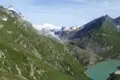 Türkisfarbener Stausee in den Alpen, umgeben von grünen Berghängen und schneebedeckten Gipfeln.