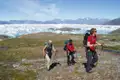 Drei Wanderer mit Trekkingausrüstung wandern durch eine Tundra-Landschaft mit Gletschern und Bergen im Hintergrund.