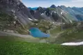 Blick auf einen blauen Bergsee inmitten der Alpen. Steile, felsige Hänge und Schneefelder umgeben den See. Eine grüne Wiese mit Wildblumen liegt im Vordergrund.