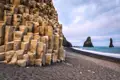Geometrische Basaltsäulenformationen am schwarzen Sandstrand mit Blick auf die charakteristischen Felsnadeln im Meer.