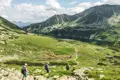 Wanderer mit Rucksäcken und Wanderstöcken auf einem Bergpfad in Richtung eines Sees und hoher Berge.