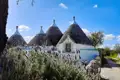Weiße Trulli-Häuser mit grauen Kegeldächern und Lavendelpflanzen im Vordergrund unter blauem Himmel.